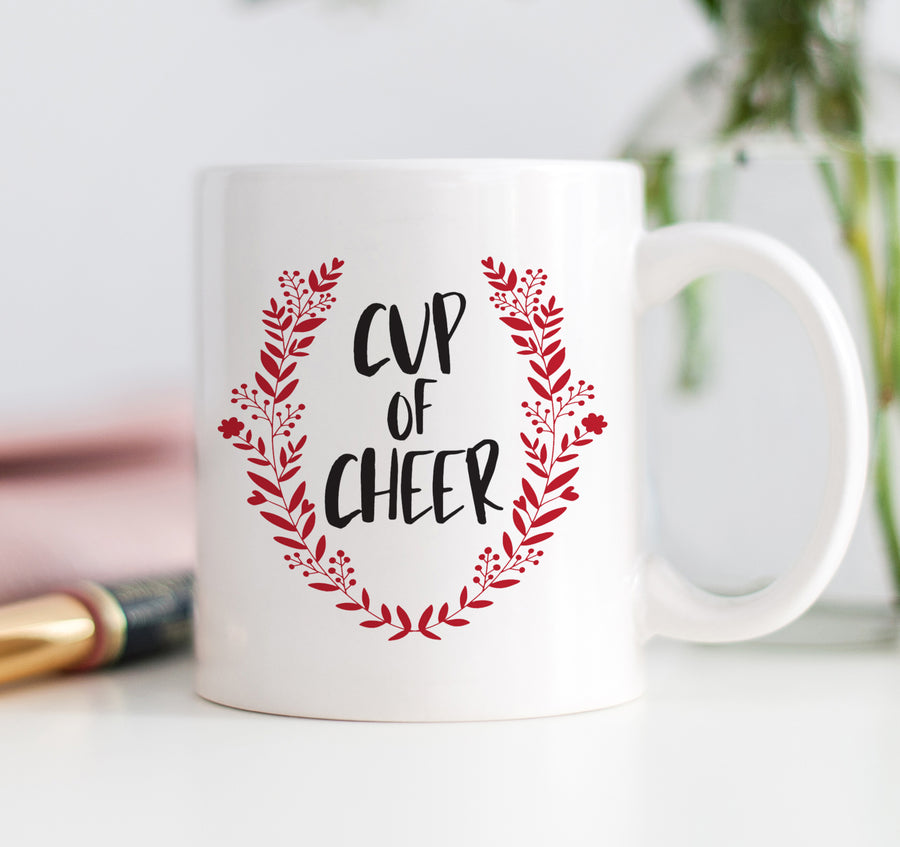 Cup of Cheer Coffee Mug with bold black festive text and a red wreath Christmas emblem on a fine white ceramic mug.