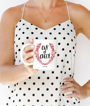 Cup of Cheer Coffee Mug with bold black festive text and a red wreath Christmas emblem on a fine white ceramic mug.